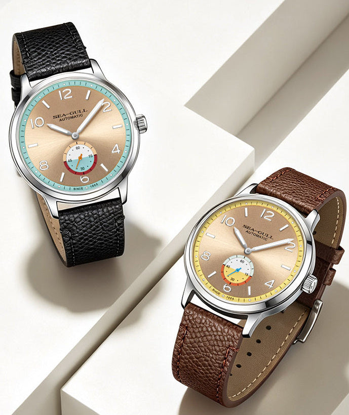 Two wristwatches displayed on white geometric blocks. One watch has a black textured leather strap and a beige dial with a light blue bezel and a small multicolor moon phase subdial.