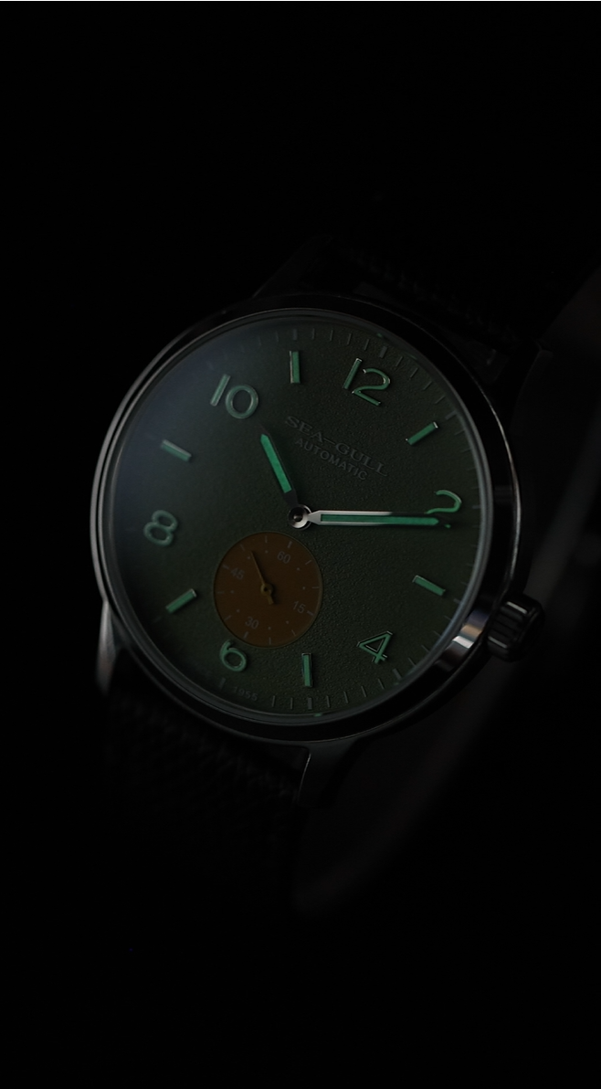 A wristwatch with a dark green dial, luminous green hands and numerals, and a small gold-toned circular subdial, shown against a black background.